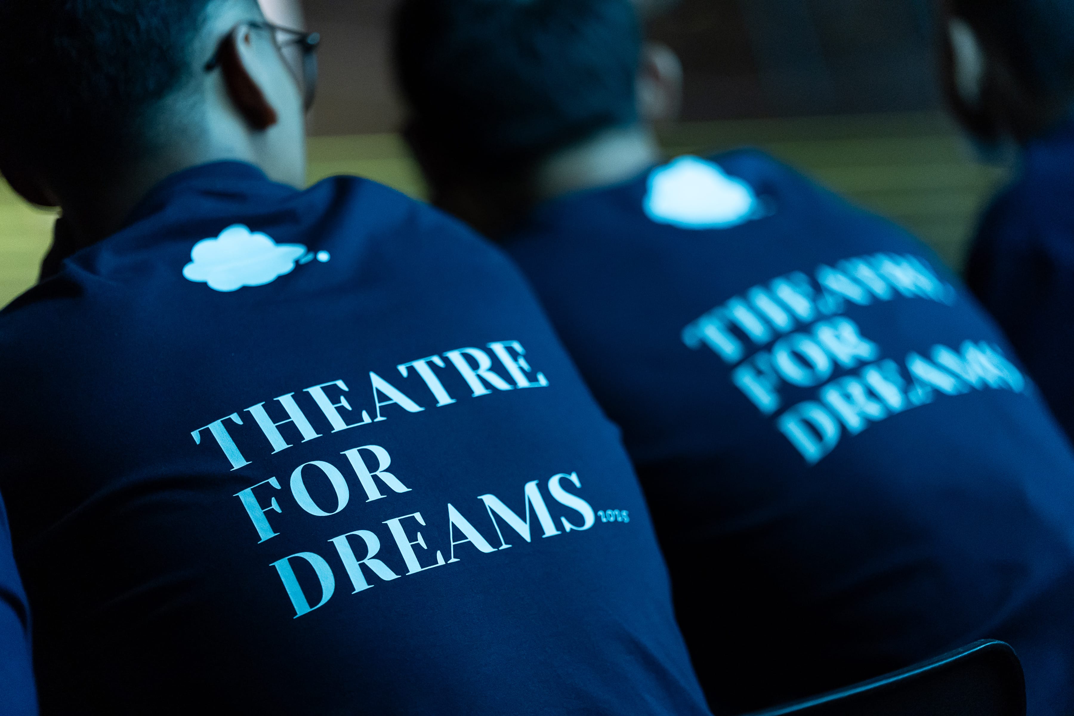Close-up of participants seated in a row, wearing navy blue 'Theatre for Dreams' t-shirts.